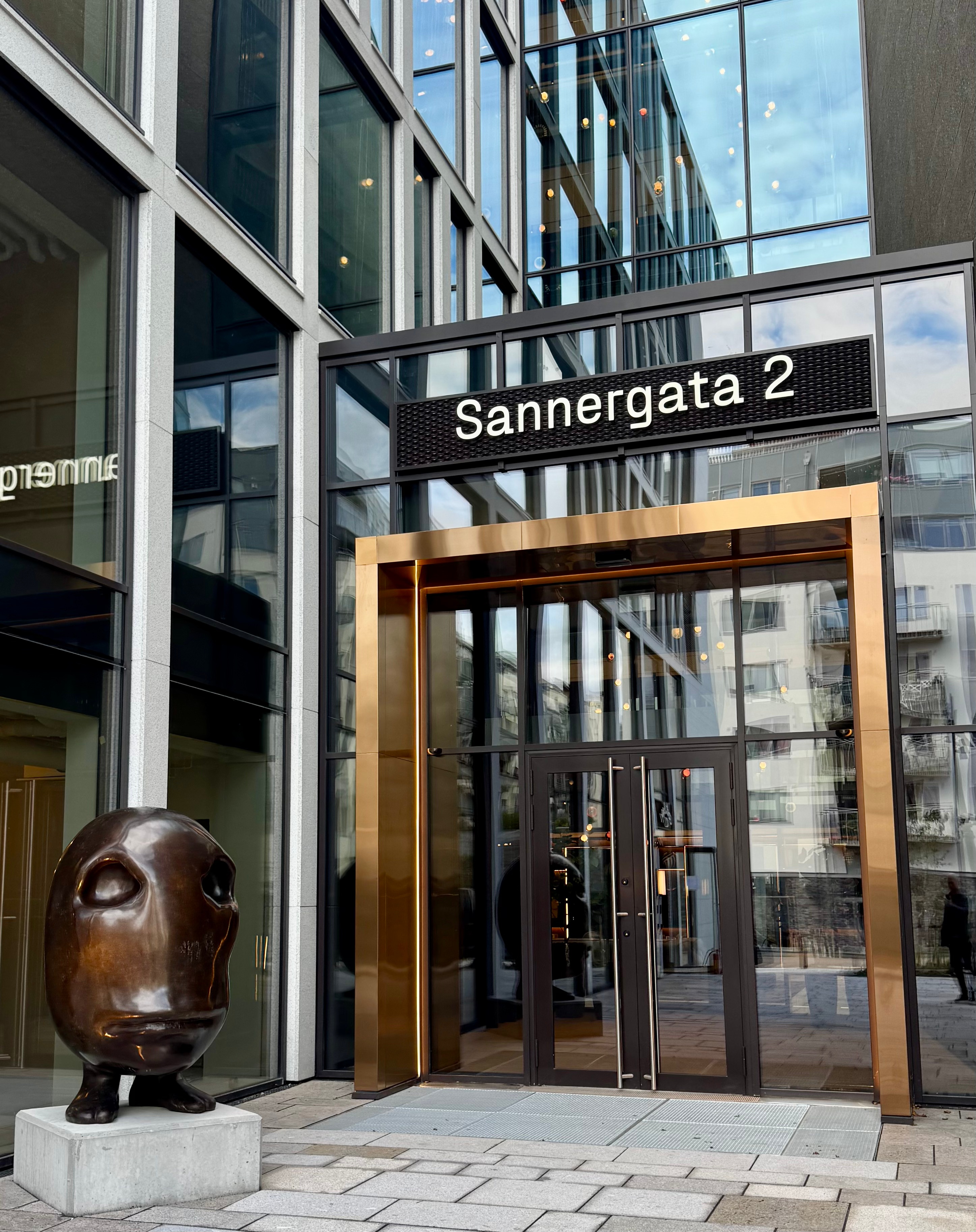 Inngangspartiet til Sannergata 2 i Oslo med moderne glassfasade, gyllen ramme rundt hovedinngangen og skulptur ved siden av døren.