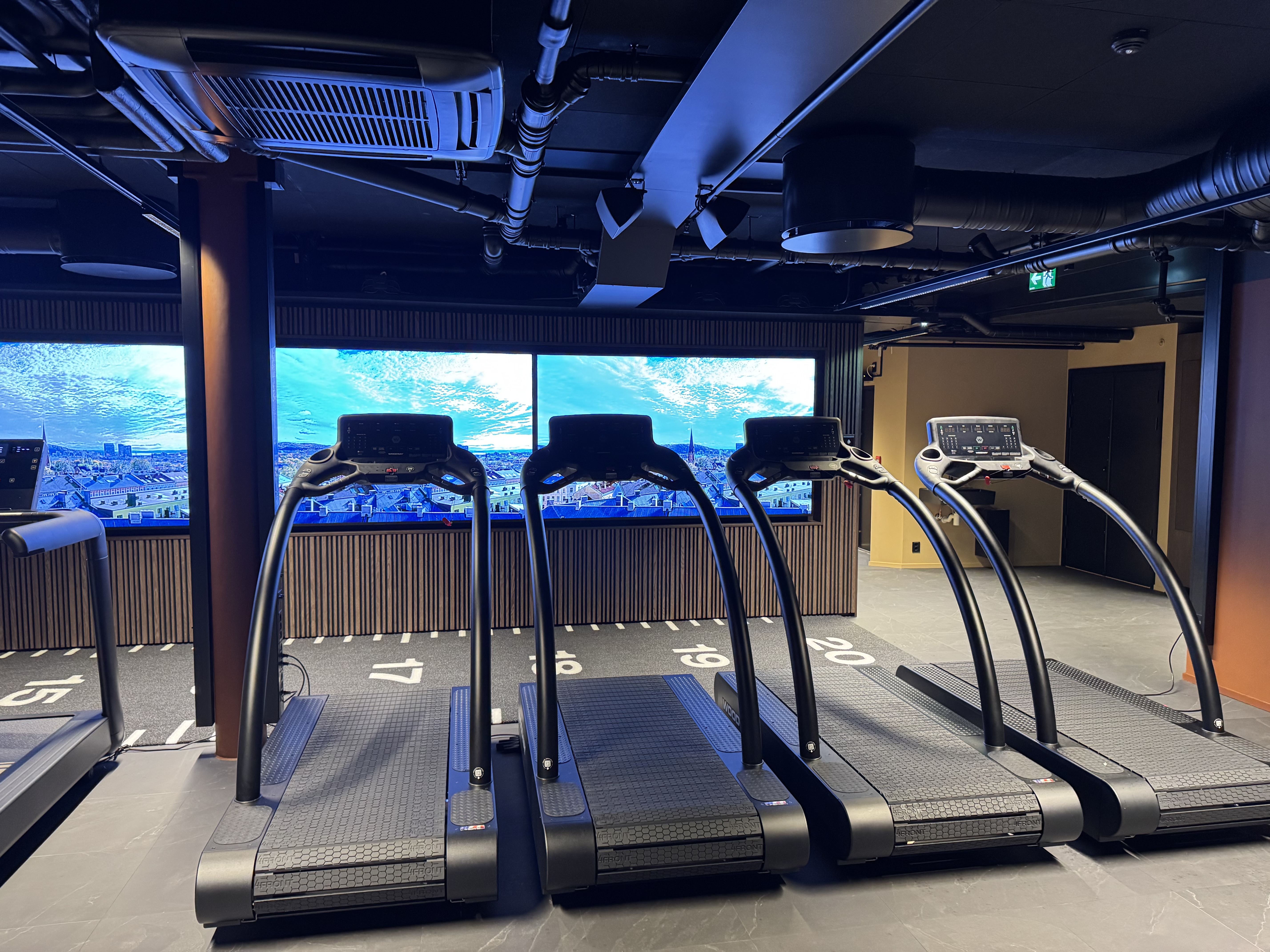The image shows a modern gym with four black treadmills lined up in a row, each with a unique design and features. The tre...
